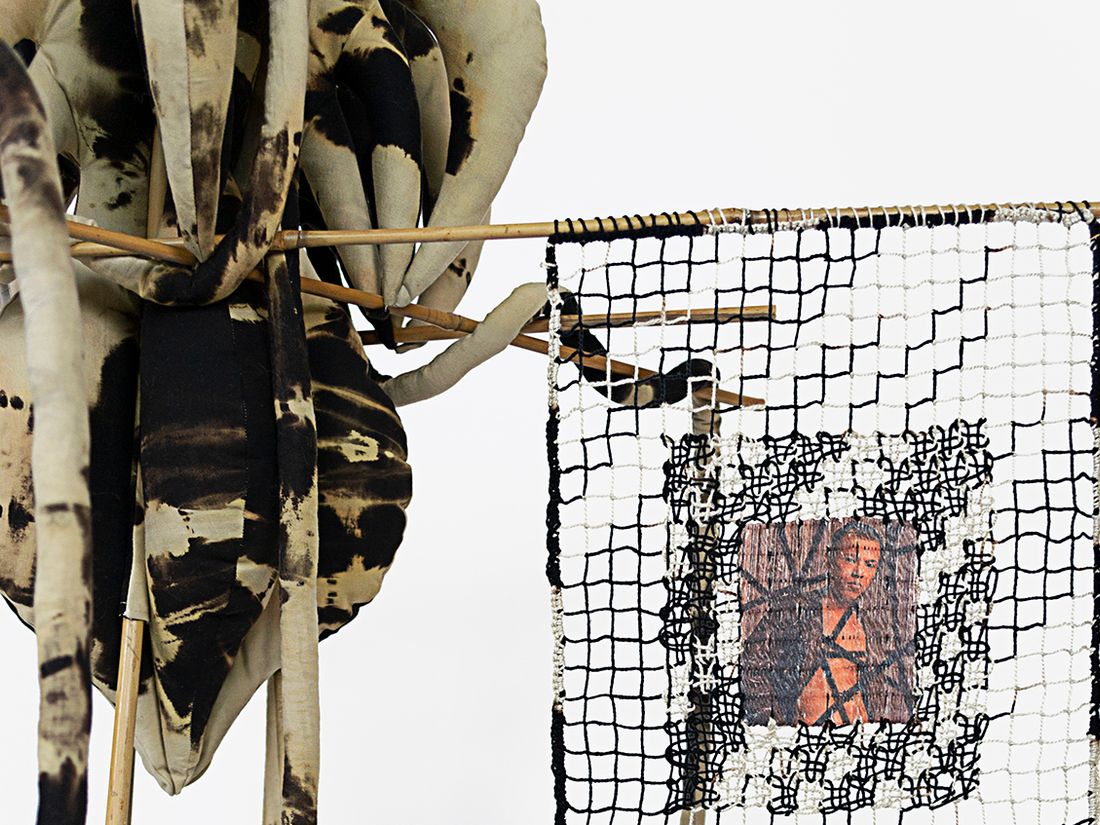A free standing soft sculpture reverse tie dyed in hues of black and beige with filet lace and netting elements framed in bamboo. Within the filet lace is an embedded gay pornographic image using a sublimation dye technique. The soft sculpture element has a main body suspended in the middle of the bamboo frame with holes where a vine-like pieces are twisted around.