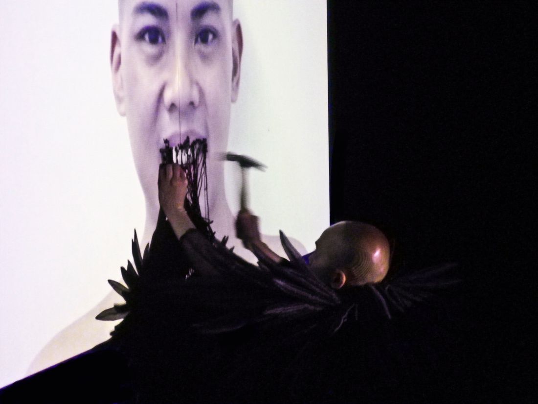 A performance of the artist, Jade Yumang, who is wearing a handmade costume while hammering and destroying a drywall with a video projection of the artist holding a smile in a black box theater. 