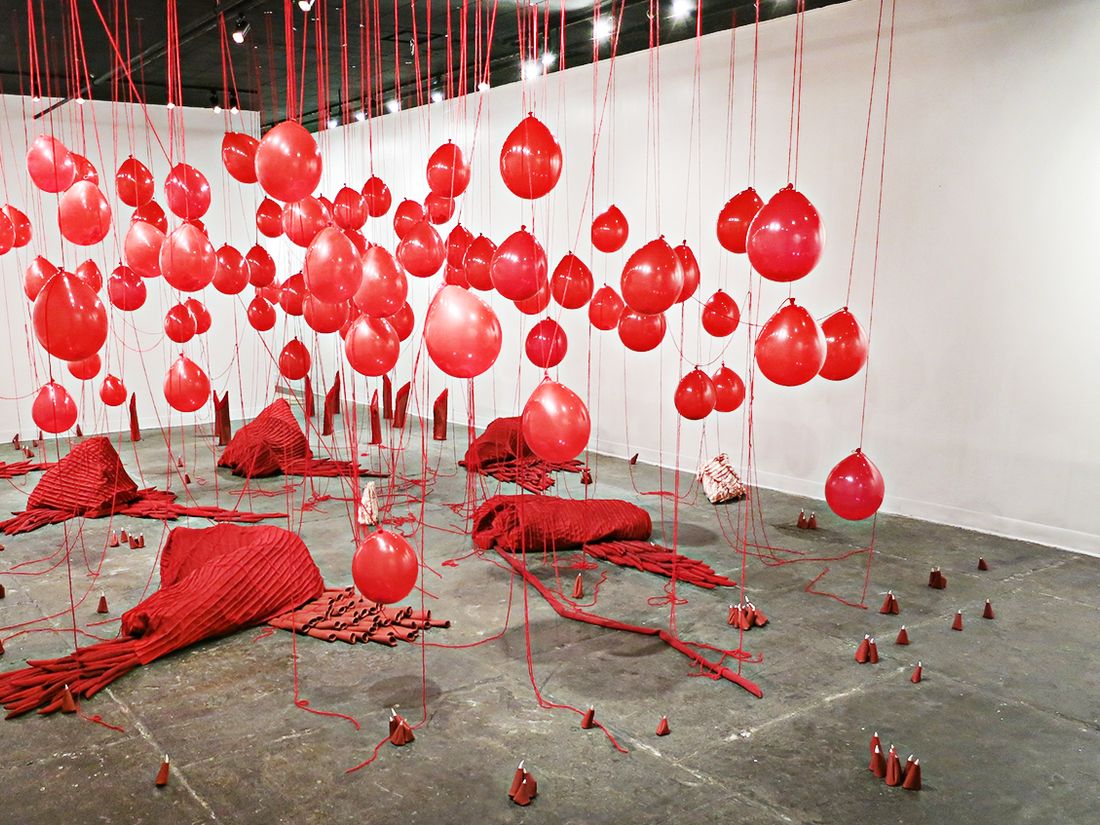 Installation shot of a show at the Leslie-Lohman Museum of Art, New York, NY, USA. The installation consists of red balloons hanging from red yarn attached to the ceiling, on the floor are soft sculptures in red with cylindrical elements made with vinyl, and there are small talon-like sculptures made with red vinyl and ceramic nails.